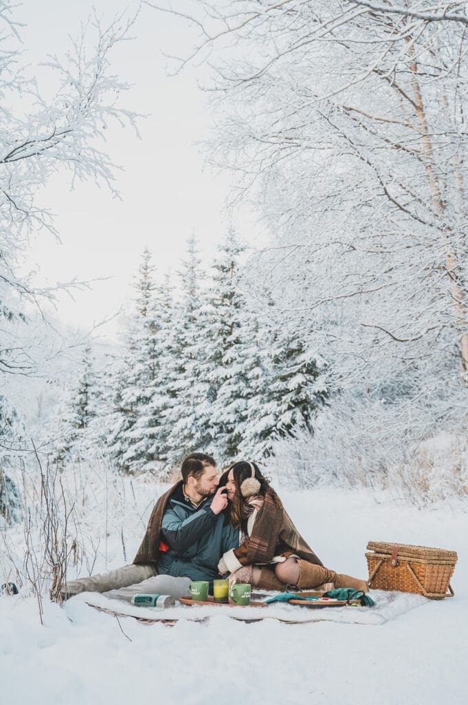 couple sitting in snow having picnic winter engagement photos Alaska