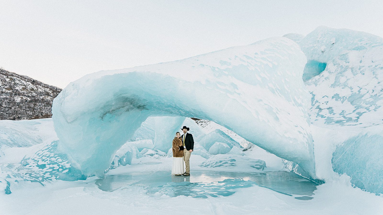 Bride and groom standing beneath a blue ice arch during their Knik Glacier elopement in Alaska