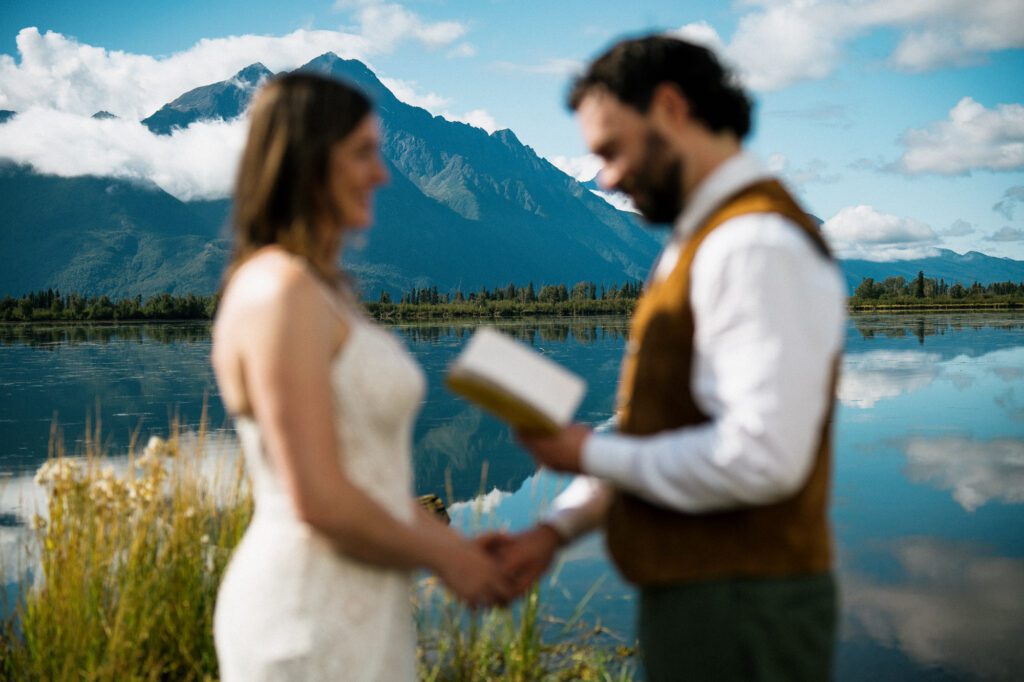 Bride and groom standing together beside an Alaska lake with mountains in the background