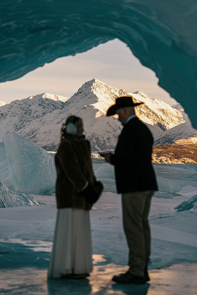 Bride and groom exploring blue ice formations at Knik Glacier with snow-covered Alaska mountains behind them